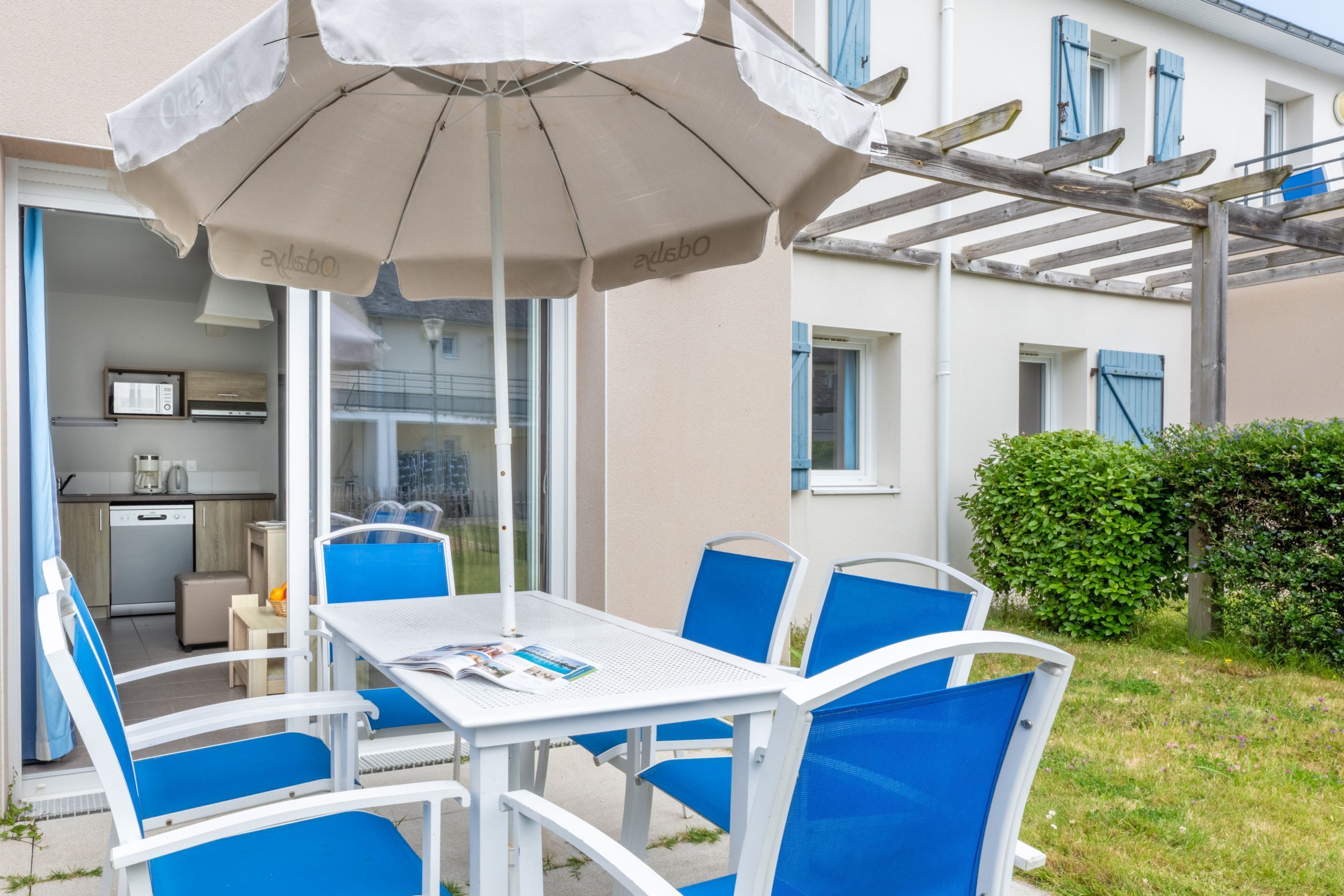 An outdoor patio with a table and chairs, a pergola, and a view of a house with a kitchen and dining area.