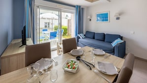 A dining area with a table set for two, a blue sofa, and a view through a glass door to an outdoor patio with chairs and umbrellas. - Résidence Odalys Les Iles du Morbihan (Baden)