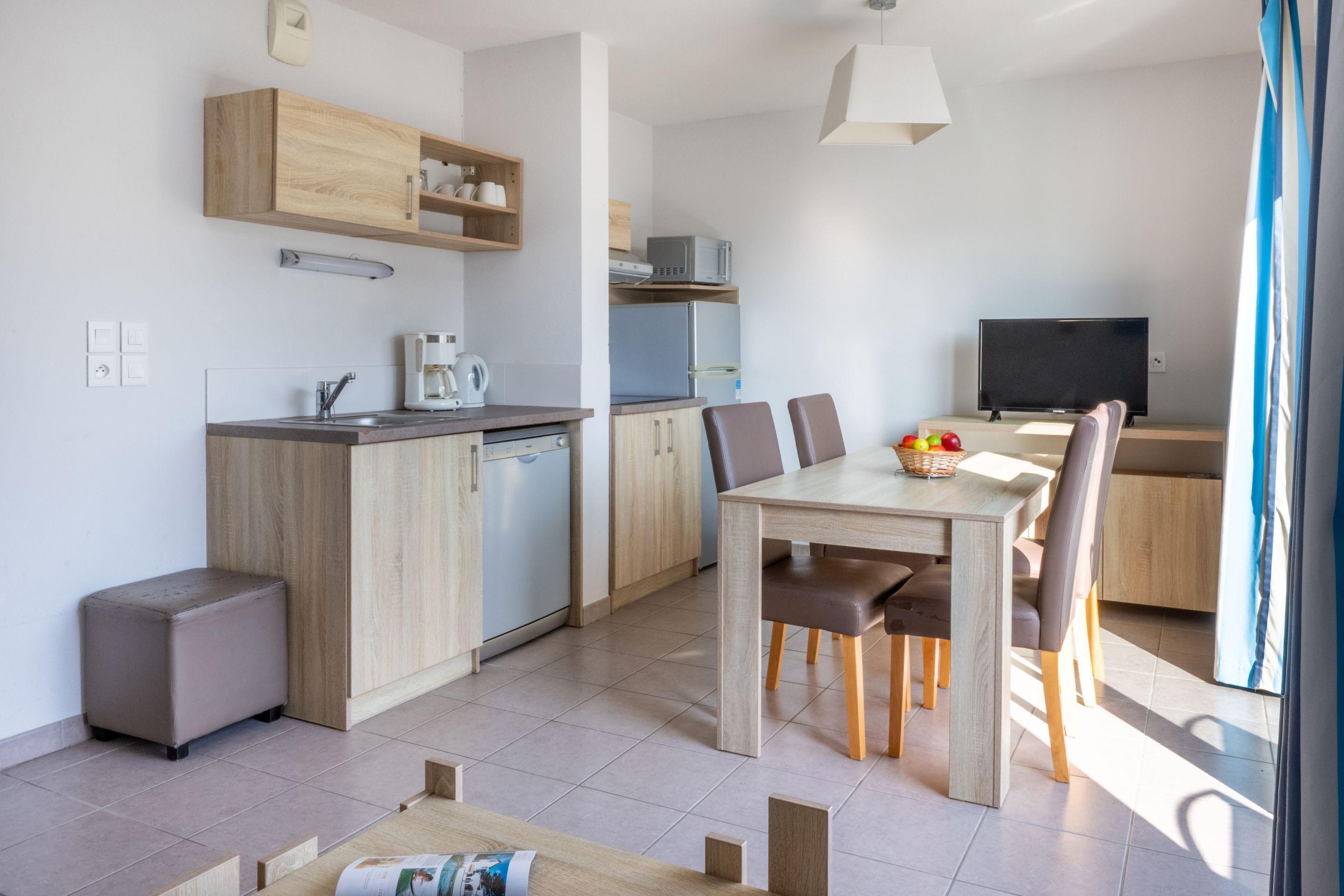 Een moderne keuken met houten kastjes, een ingebouwde magnetron en een koffiezetapparaat. Er is een eethoek met een tafel en stoelen, en een televisie op de achtergrond.