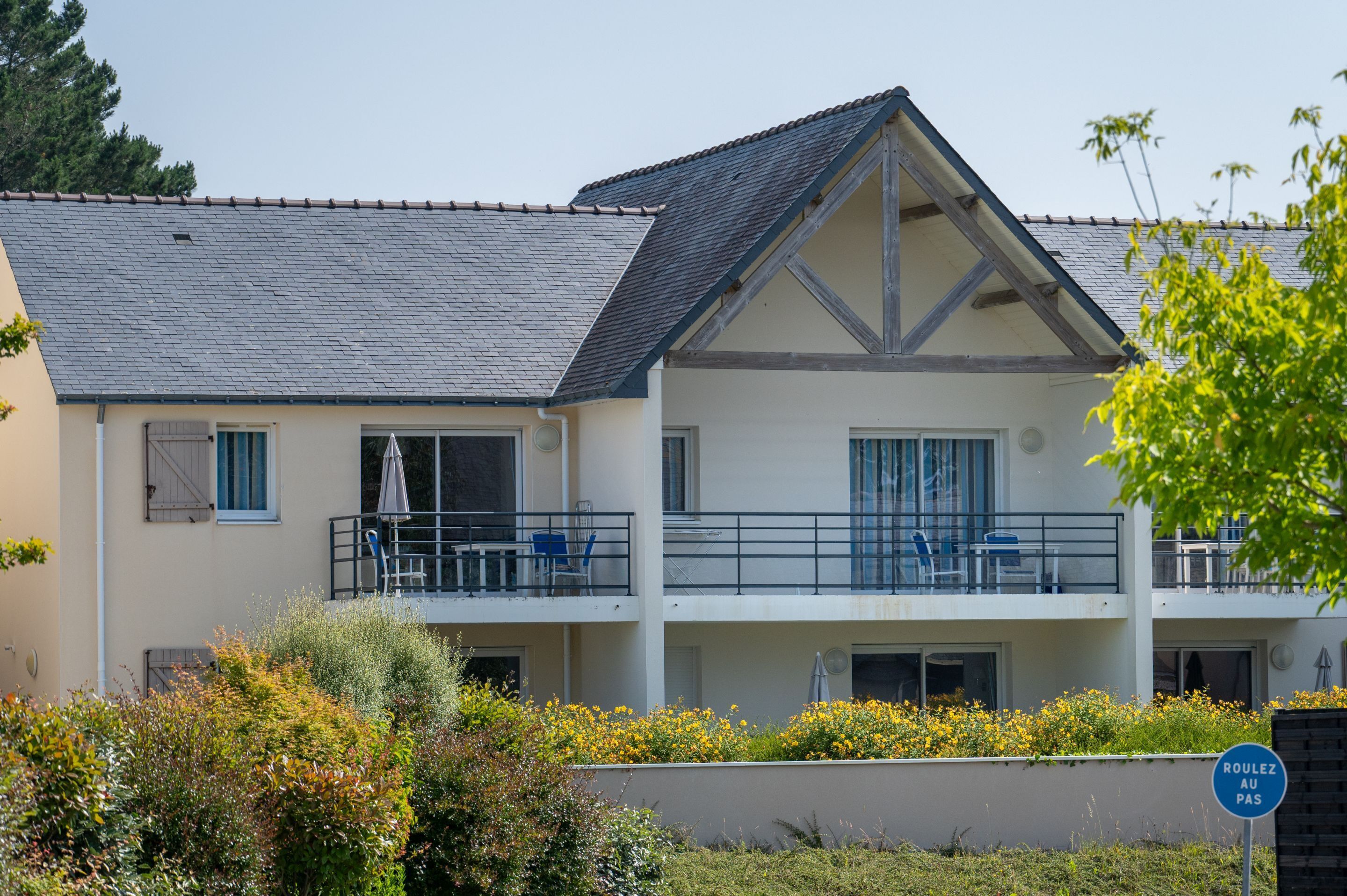 Een gebouw met twee verdiepingen, een balkon en een tuin op de voorgrond. Op het bord staat 