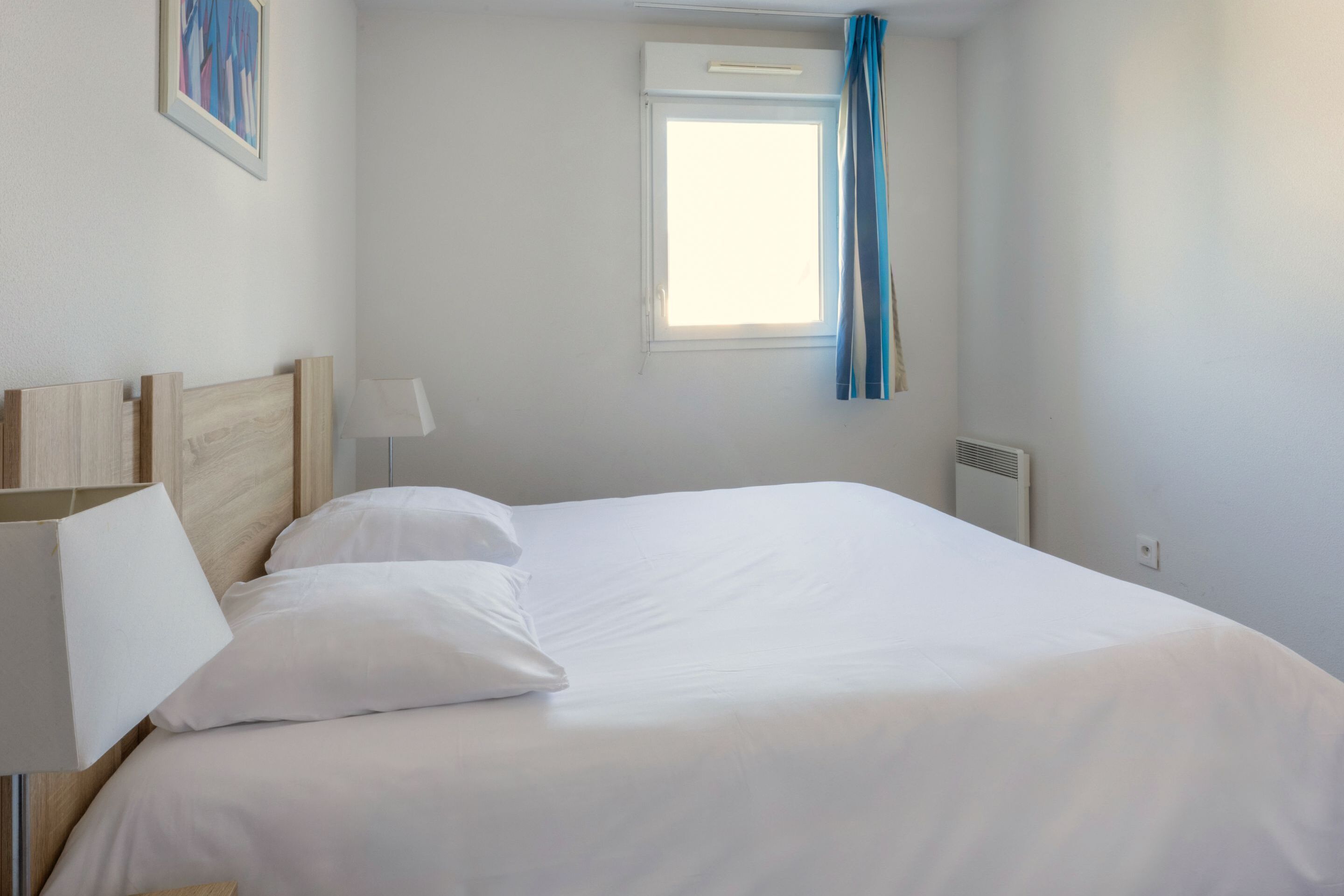 Ein ordentlich bezogenes Bett mit weißen Leinen, ein hölzernes Kopfteil, eine Nachttischlampe und ein Fenster mit blauen Vorhängen.