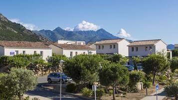 A row of white buildings with red-tiled roofs, a parked car, and a mountain backdrop.