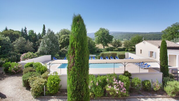 A pool area with lounge chairs and a tall tree in the foreground. - Résidence Odalys Les Soleils du Luberon (Bonnieux)