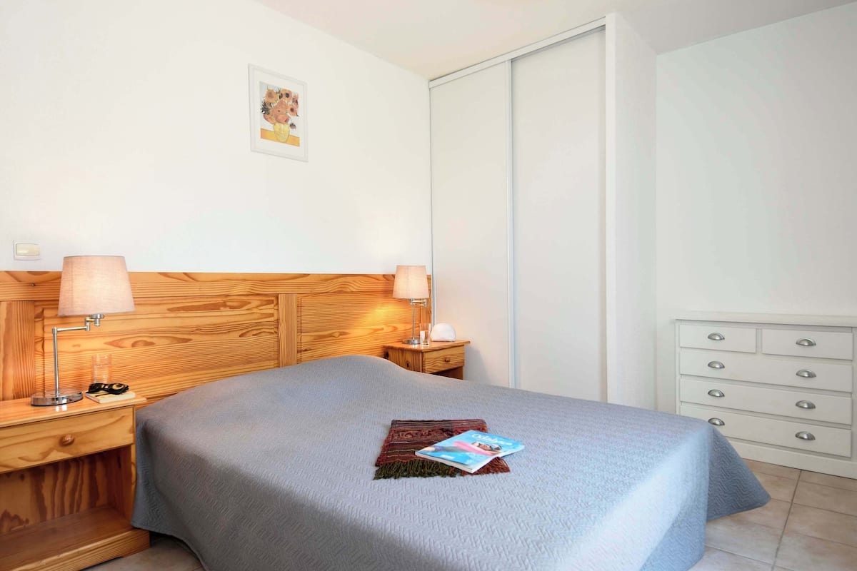A neatly made bed with a grey bedspread, two bedside lamps, a wooden headboard, a white dresser with drawers, and a framed picture on the wall.