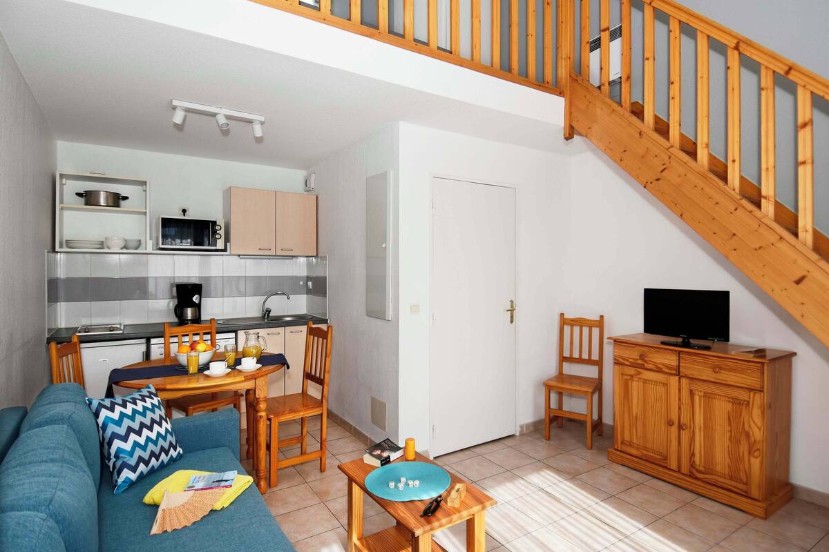 A compact living space with a kitchenette, a dining area, a sofa, a TV stand, and a wooden staircase.