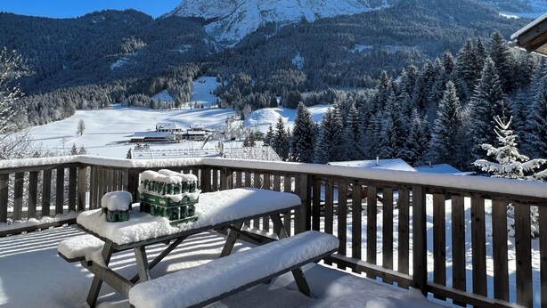 Une terrasse en bois avec un banc et une table de pique-nique recouverts de neige, offrant une vue sur un paysage montagneux enneigé sous un ciel dégagé.
