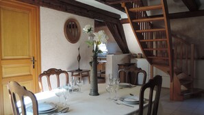 Un comedor con una mesa redonda dispuesta para cuatro personas, sillas de madera, un florero con flores y una escalera de madera al fondo.
