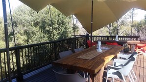 Um pátio externo com mesa de madeira e cadeiras brancas, cercado por um guarda-corpo de madeira e coberto por um toldo grande.
