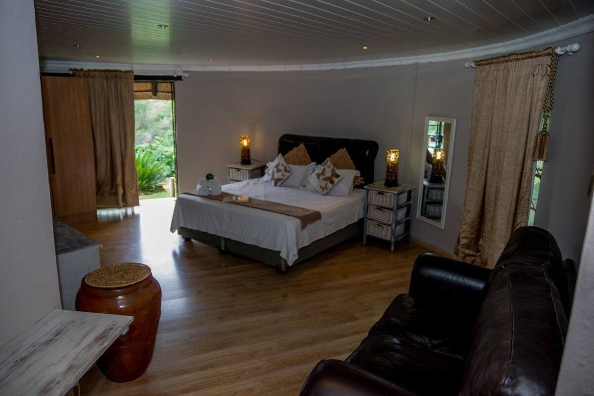 A bedroom with a bed, bedside tables, a wooden floor, a leather sofa, and a view of greenery outside.