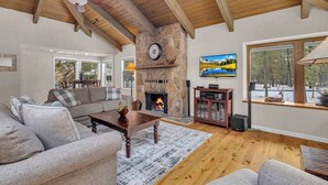 A cozy living room with a stone fireplace, a large window with a view of trees, and a ceiling fan. - Unique spacious cabin with private deck, hot tub! (Sunriver)