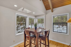 A dining area with a wooden table and chairs, large windows with a view of a snowy landscape, and a ceiling with wooden beams.