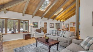 A spacious living room with a high ceiling, wooden beams, and large windows offering a view of the outdoors. - Unique spacious cabin with private deck, hot tub! (Sunriver)