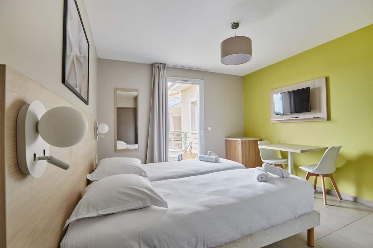 Quarto de hotel com duas camas, uma escrivaninha, uma cadeira, uma televisão e uma janela com cortinas.