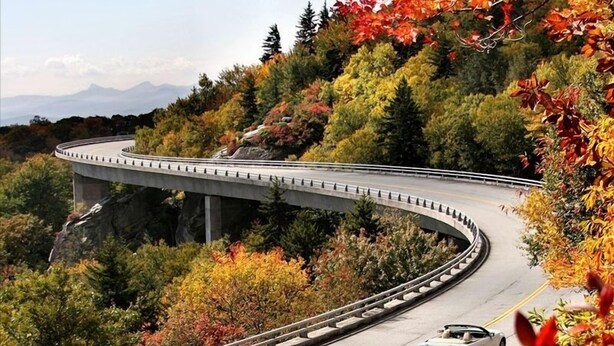 Une route sinueuse avec un pont enjambant une falaise rocheuse, le tout entouré d’un feuillage automnal.