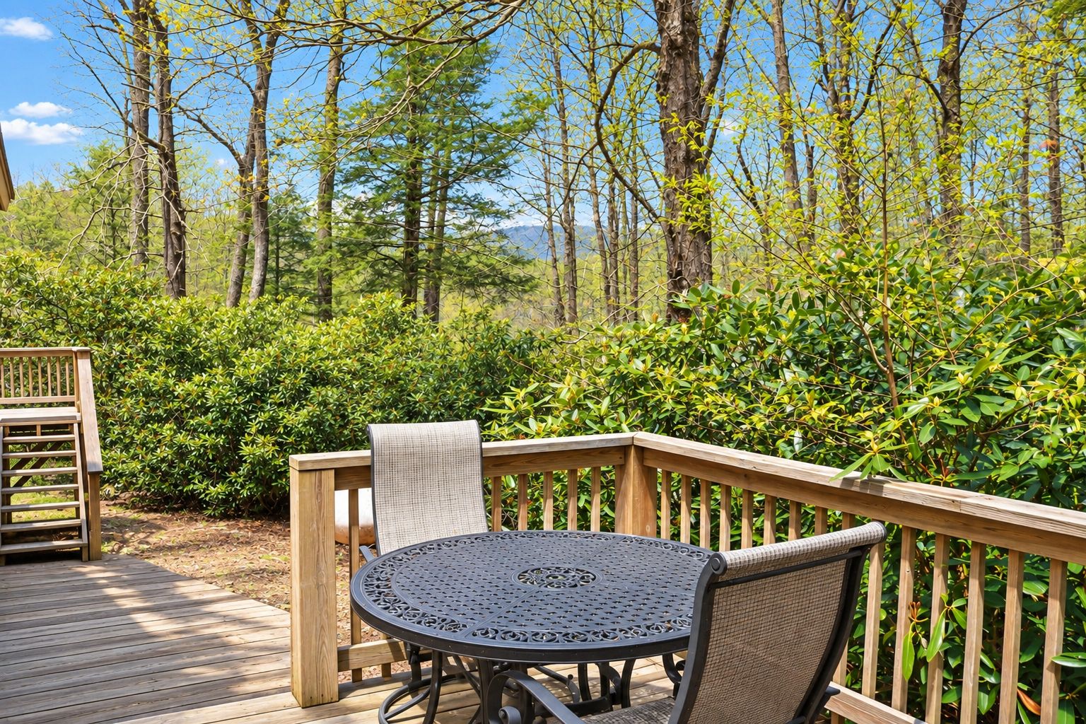 A wooden deck with a round metal table and chairs, surrounded by dense greenery and trees.