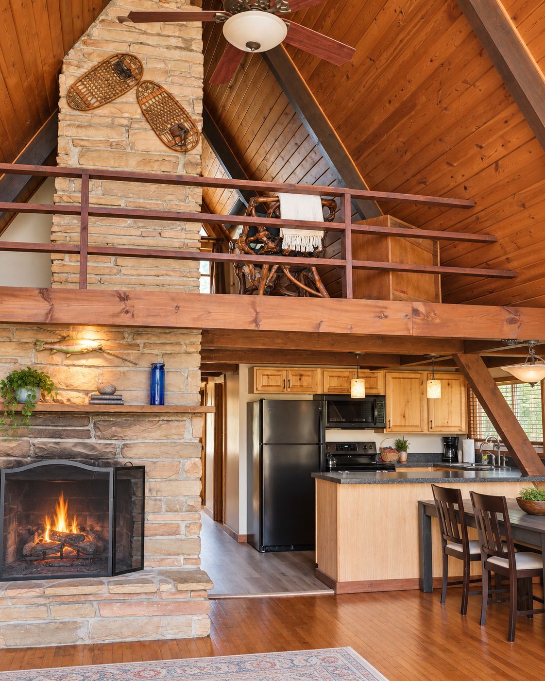 A rustic cabin interior with a stone fireplace, wooden ceiling beams, a kitchen area, and a dining space.