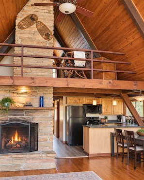 A rustic cabin interior with a stone fireplace, wooden ceiling beams, a kitchen area, and a dining space.