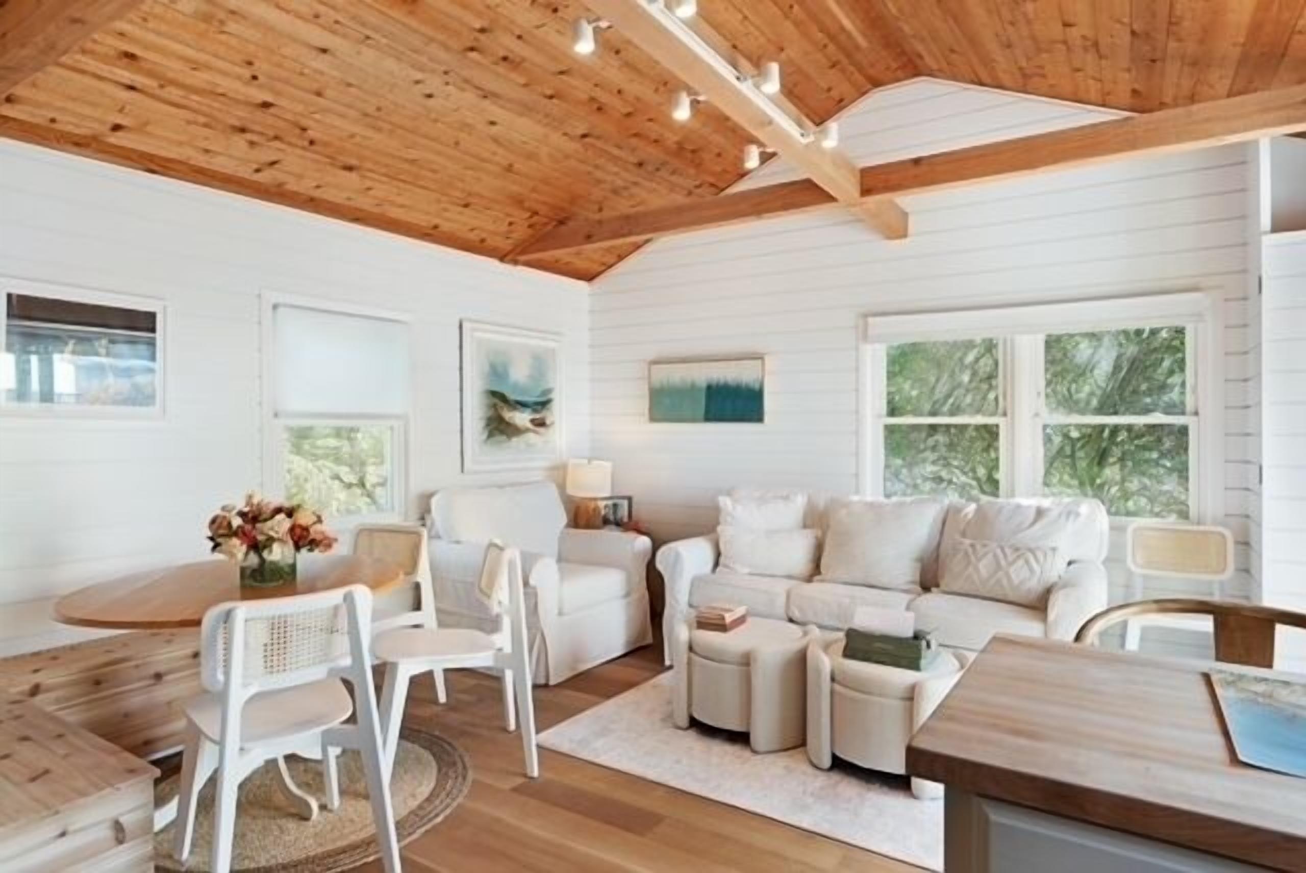 A cozy living room with a wooden ceiling, white walls, and a large window. There is a white sofa, a dining table with chairs, and a coffee table.