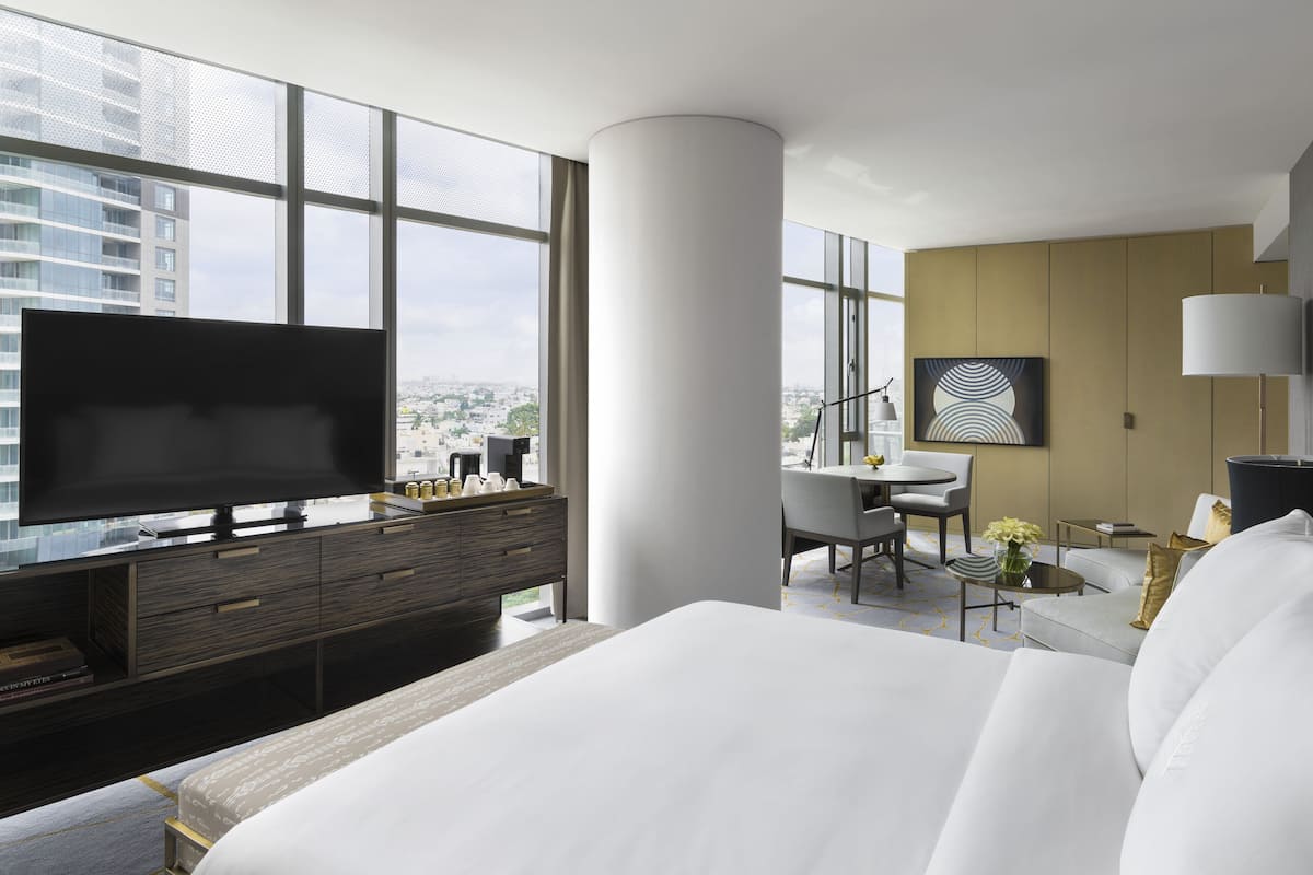 A modern hotel room with a large bed, a flat-screen TV, a desk with a chair, and a view of the city through large windows.