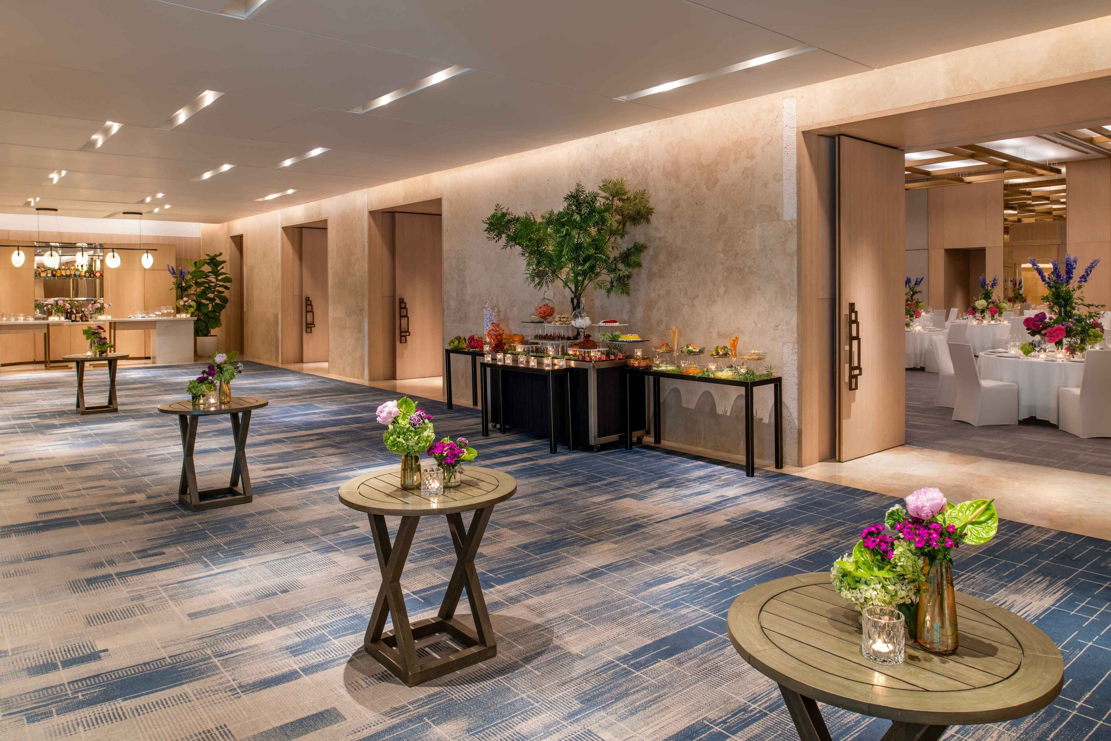 Un vestíbulo de hotel espacioso con una zona de buffet central, varias mesas adornadas con centros de flores y un diseño de techo moderno.