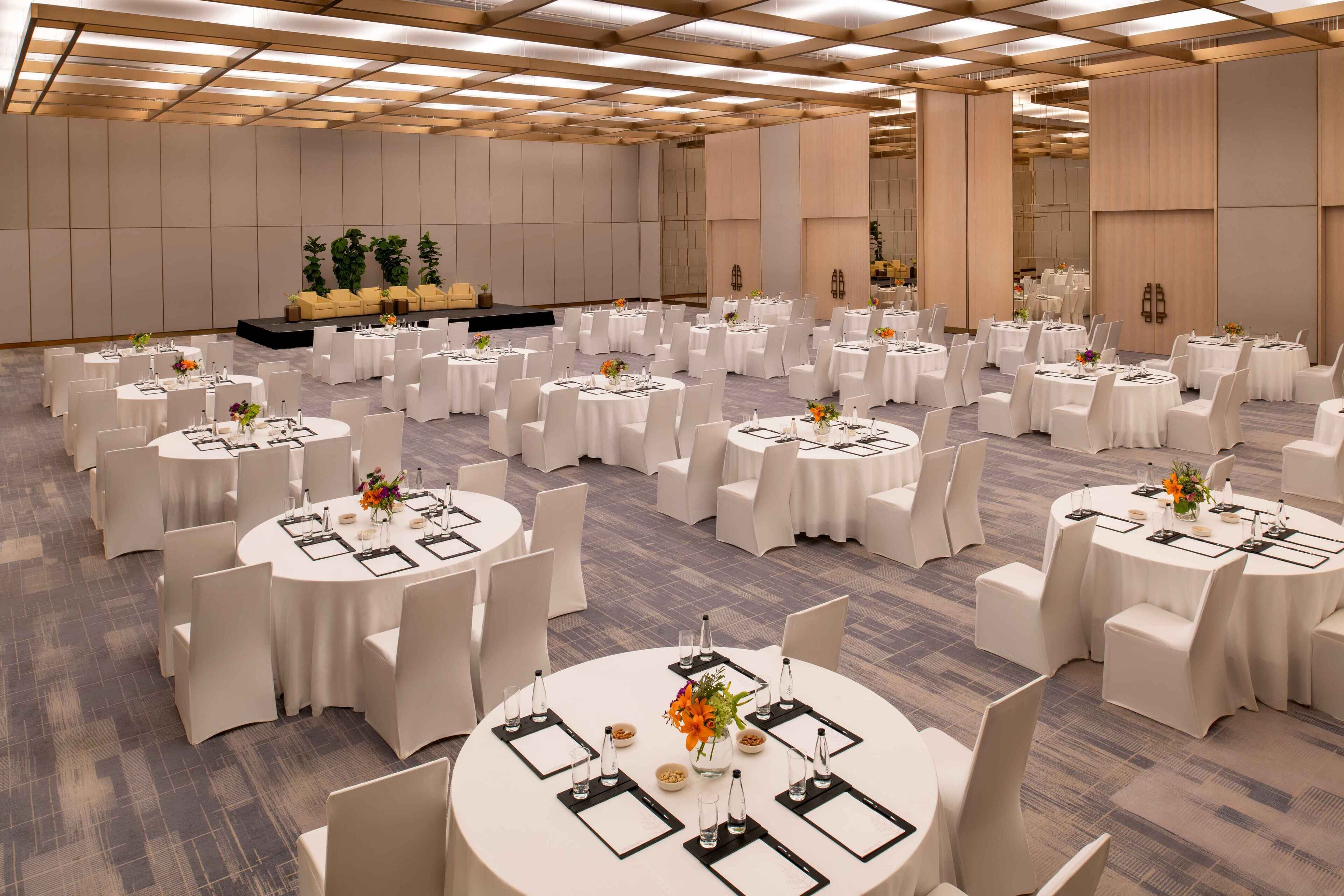 A large banquet hall with round tables set for dining, white chairs, and a stage area with plants.