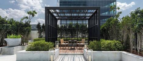 An outdoor patio area with a wooden table and chairs, surrounded by greenery and a modern building in the background.