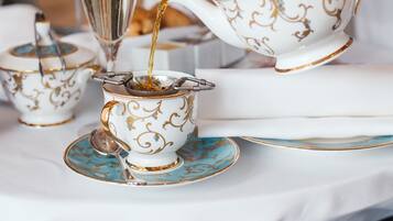 Un juego de té con tetera y tazas sobre una mesa, un pastelero con postres al fondo, y una persona sirviendo té.