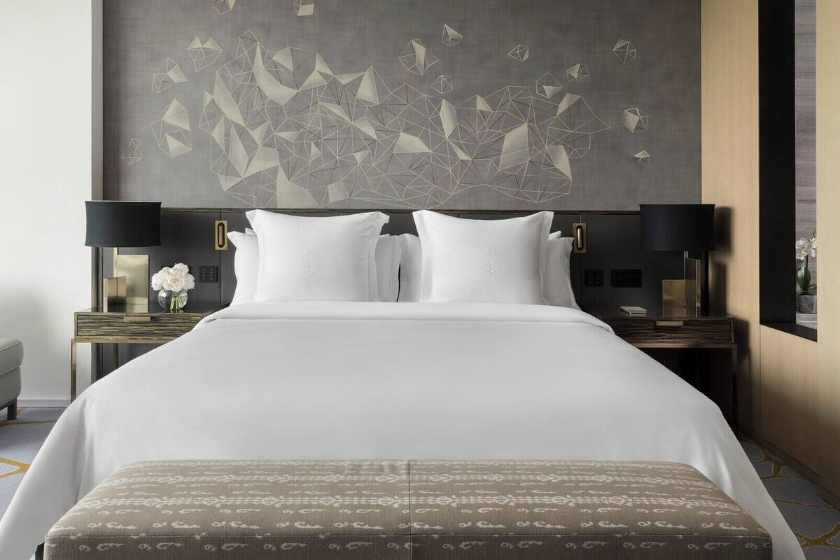 A modern hotel room with a large bed, two bedside tables, a wall art piece, and a view through a window.