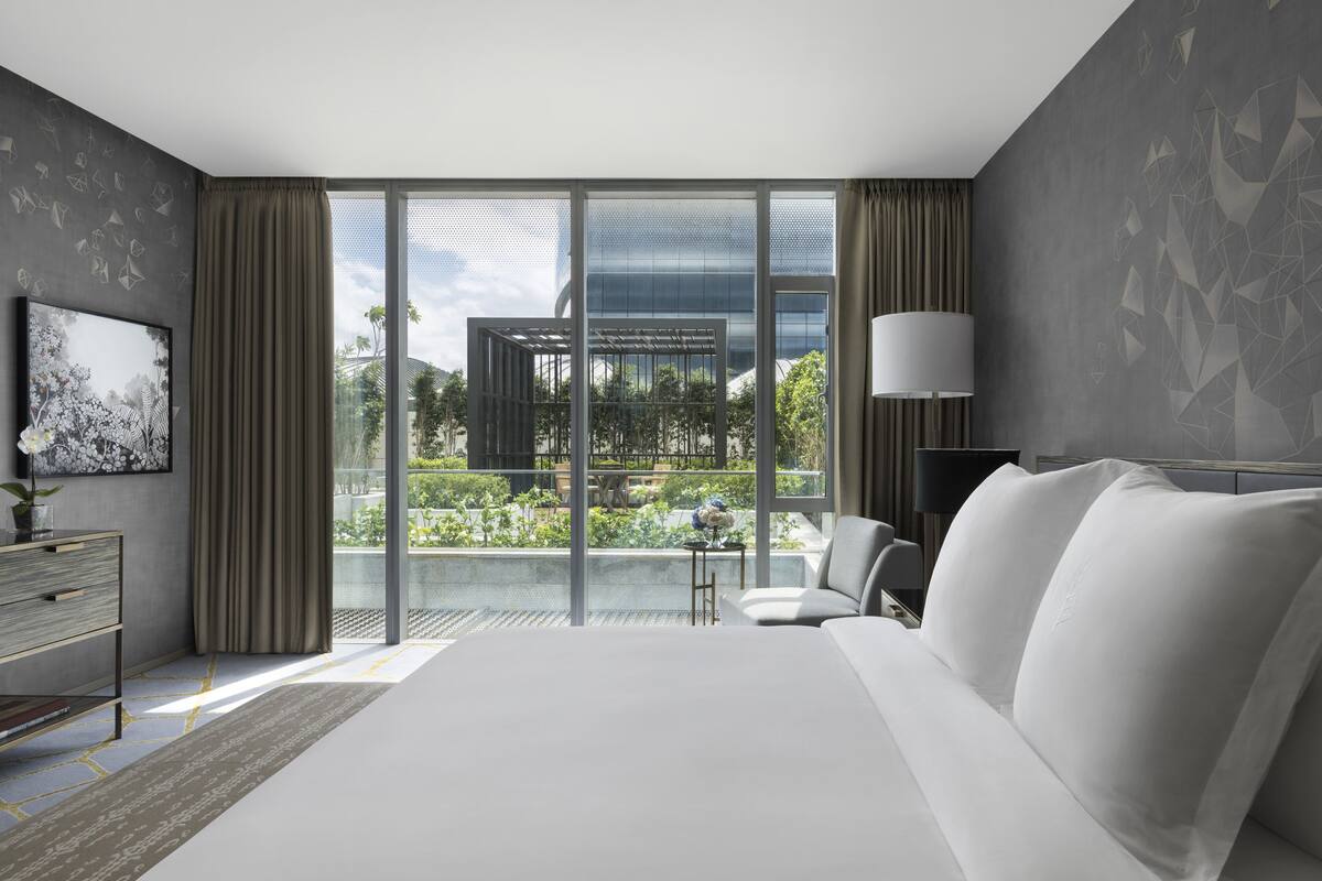 A modern hotel room with a large bed, a desk with a lamp, and a view of the outdoors through large windows.