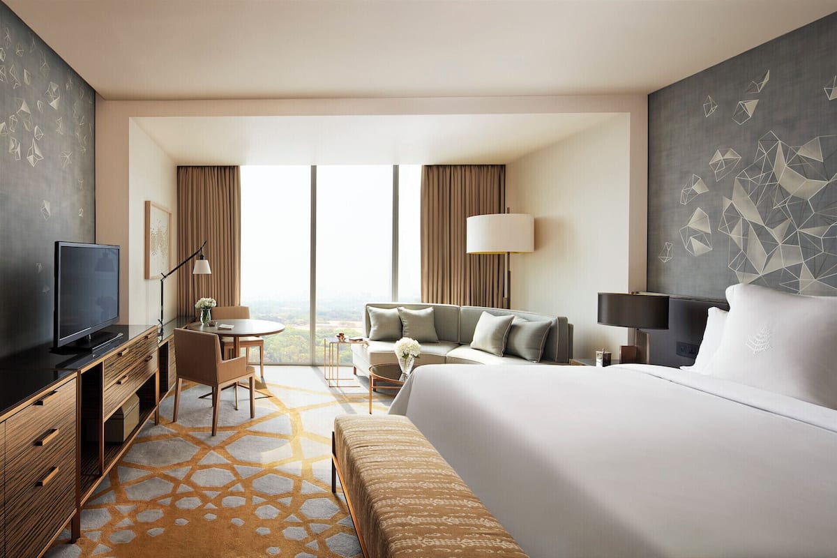 A modern hotel room with a large bed, a desk with a flat-screen TV, a sofa, and a view of the city through large windows.