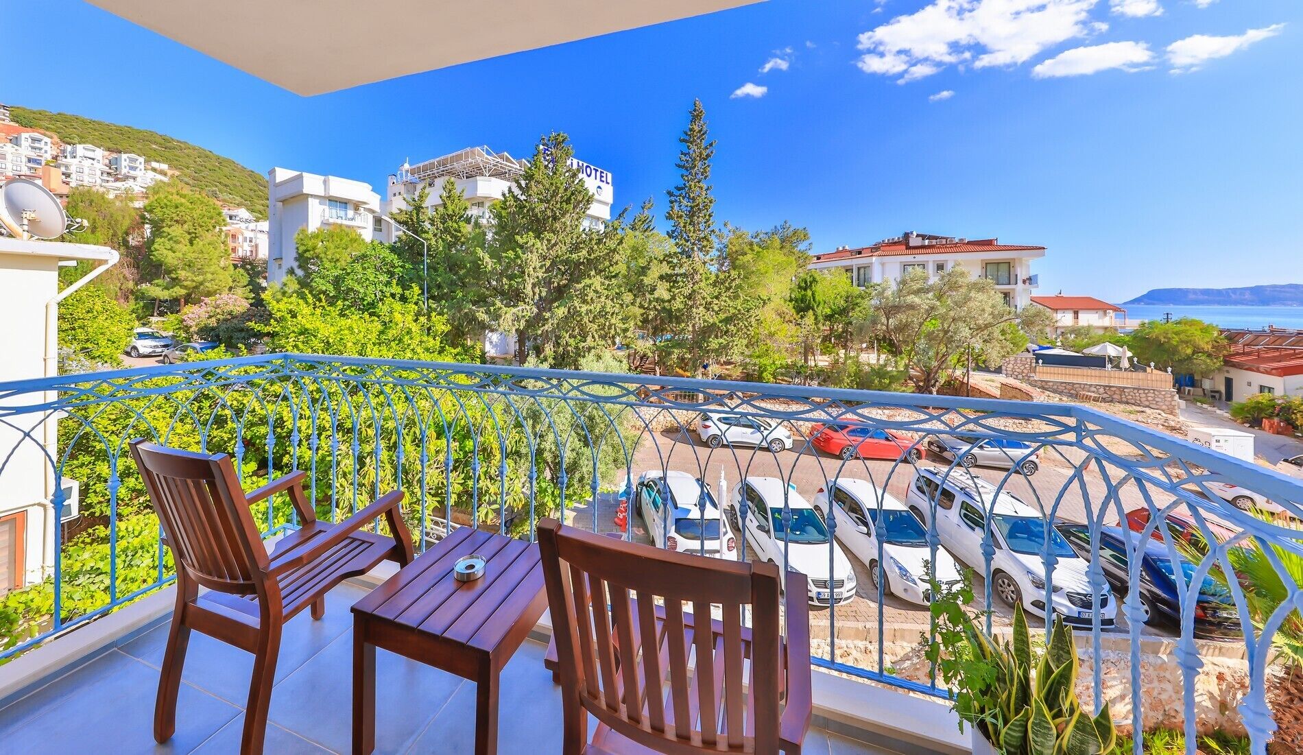 A balcony with two wooden chairs and a table, overlooking a parking area with several cars and a view of a hillside with buildings.