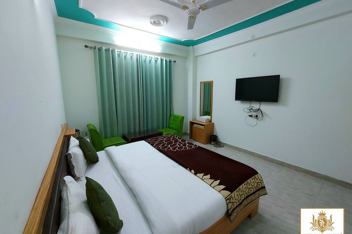 Quarto de hotel com cama, cadeiras verdes, televisão e ventoinha no teto.