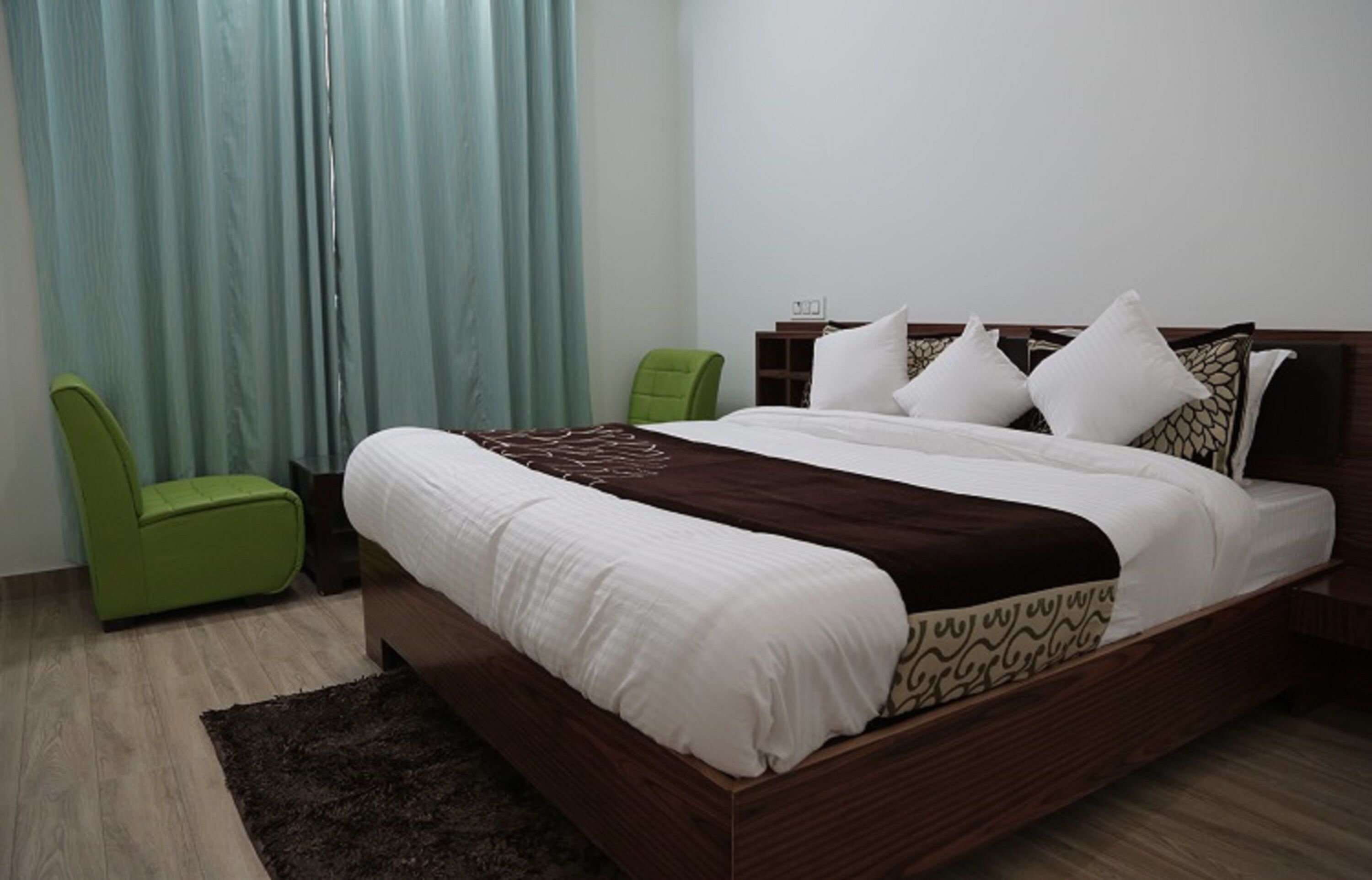 A neatly made bed with white and patterned bedding, a green chair, and a dark rug in a room with light wooden flooring and a curtain.