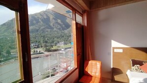 A room with a large window offering a view of a mountainous landscape and a road. - Hotel Sarla Regency (Bhuntar)