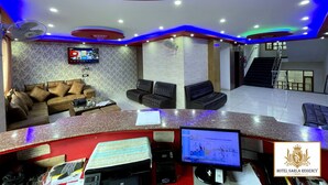 A hotel reception area with a modern design, including a curved reception desk, seating area with sofas, a flat-screen TV, and a staircase leading to an upper floor. - Hotel Sarla Regency (Bhuntar)
