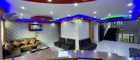 A hotel reception area with a modern design, including a curved reception desk, seating area with sofas, a flat-screen TV, and a staircase leading to an upper floor.