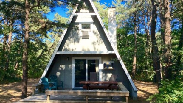 A triangular wooden cabin with a bench and chairs outside, surrounded by trees. - Cottages at the Even'tide Resort (Wellfleet)