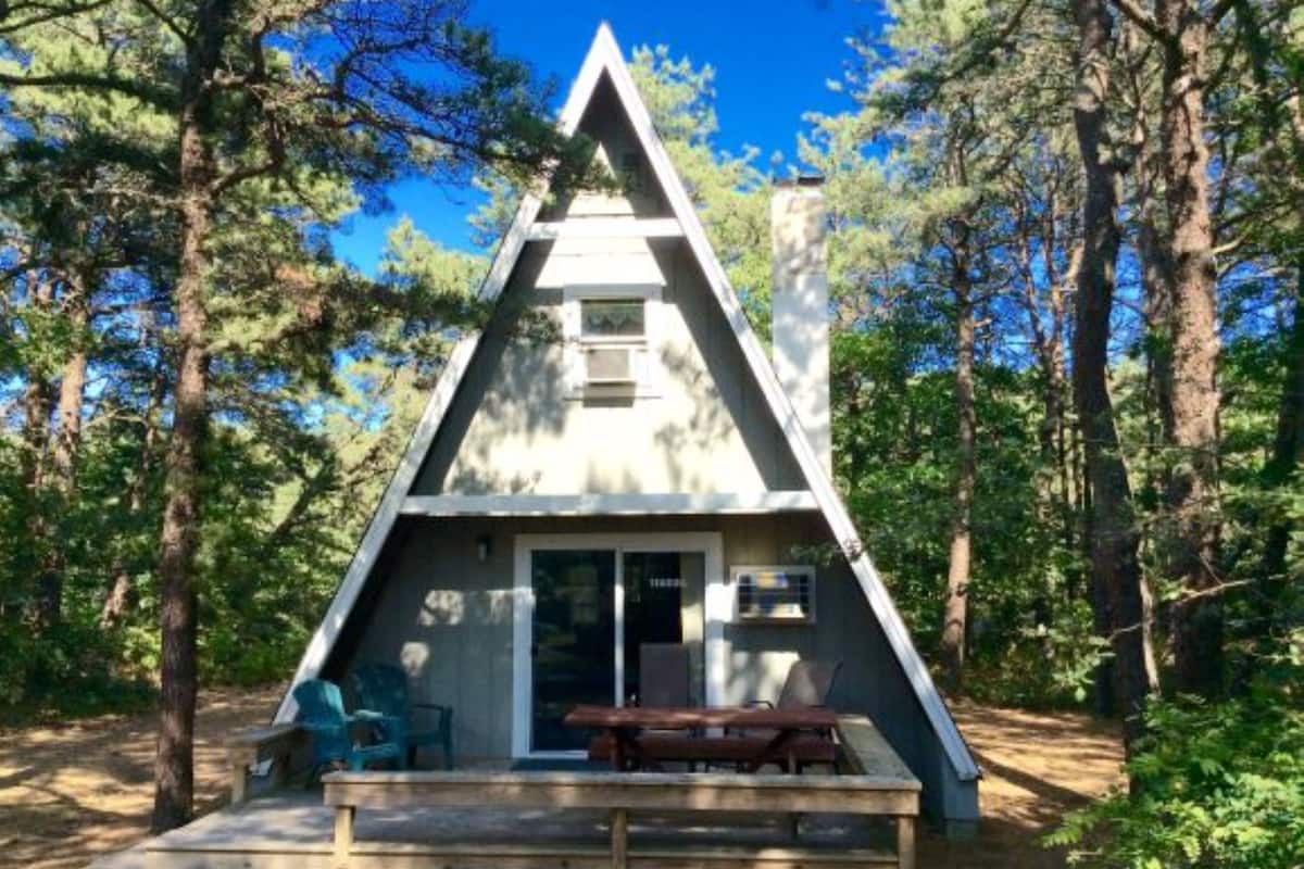 A triangular wooden cabin with a bench and chairs outside, surrounded by trees.