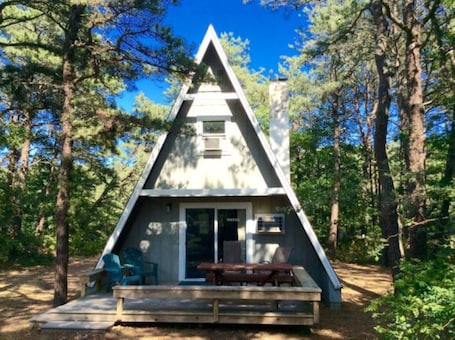 A triangular wooden cabin with a bench and chairs outside, surrounded by trees.