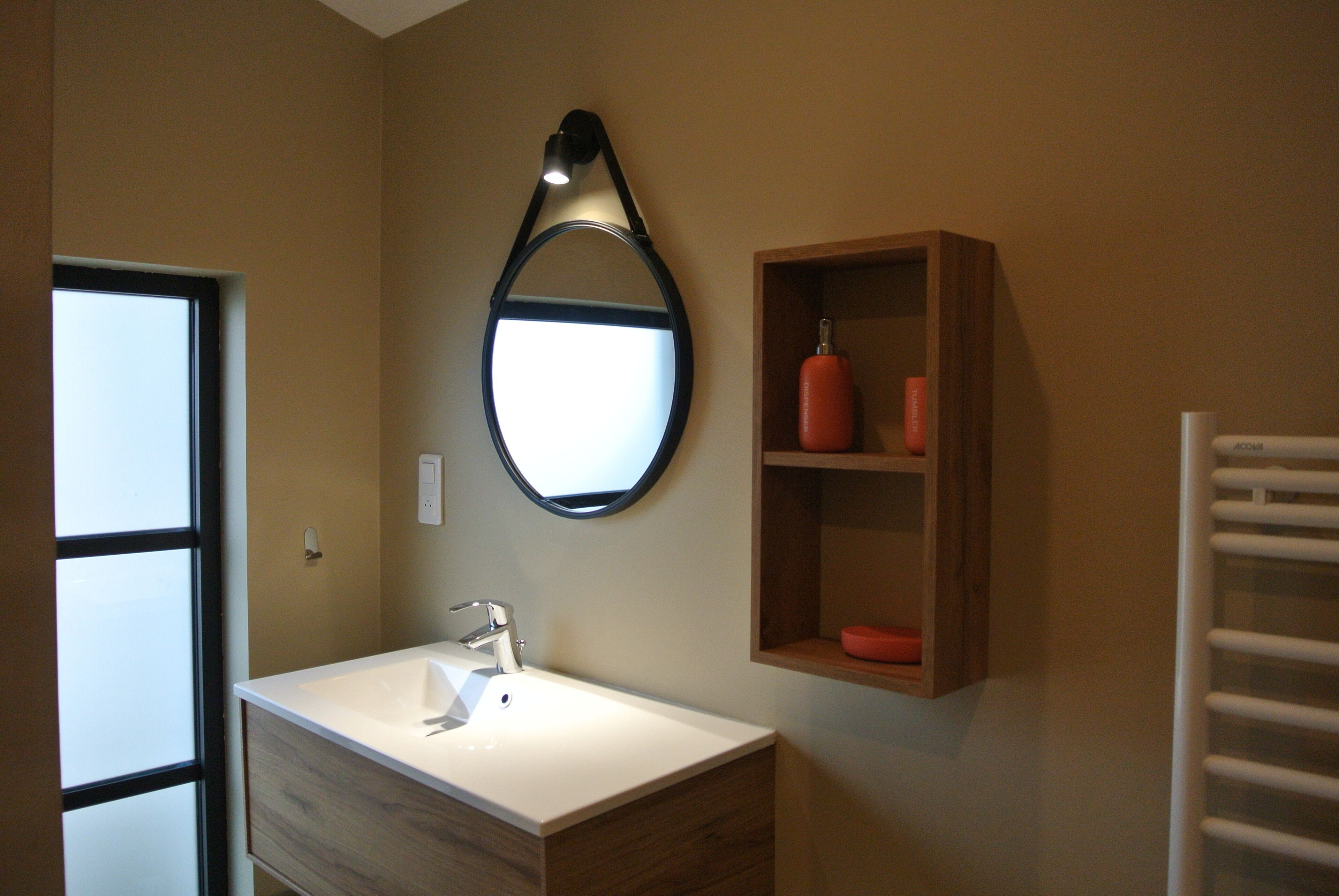 A bathroom with a white sink, a round mirror, and a wooden shelf with a red object.