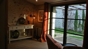 Una habitación con piso de madera, una consola blanca con objetos decorativos, un sillón estampado y un ventanal con vistas a una pared de piedra y vegetación al exterior.