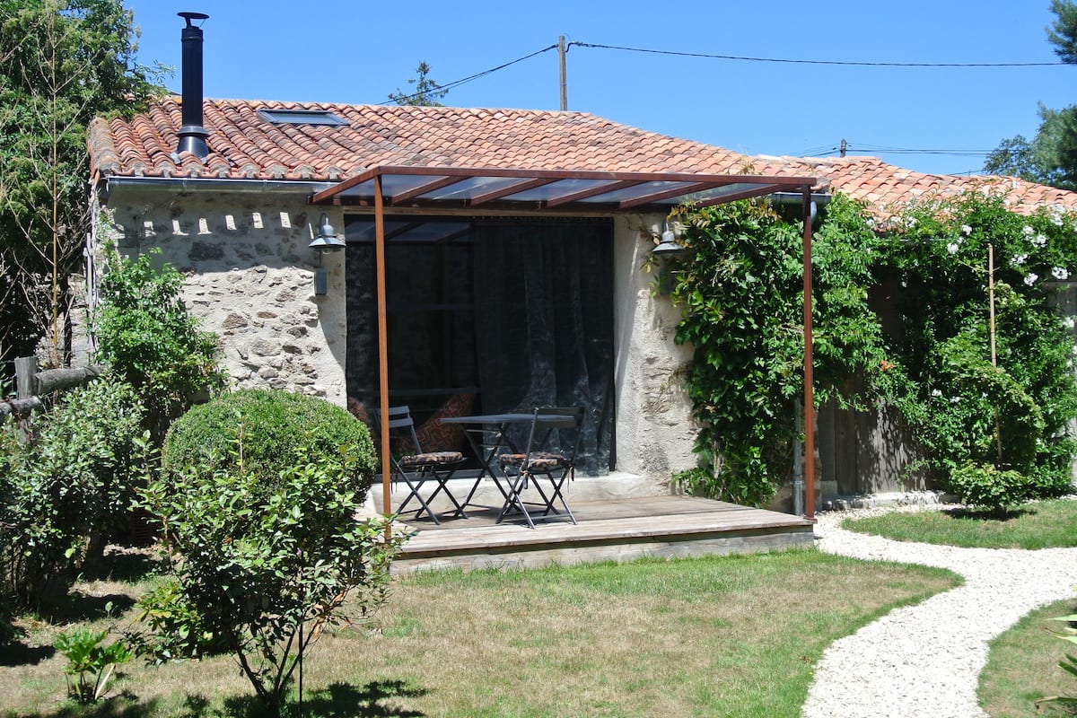 A stone building with a tiled roof, a covered patio with a table and chairs, and a gravel path leading to the entrance.