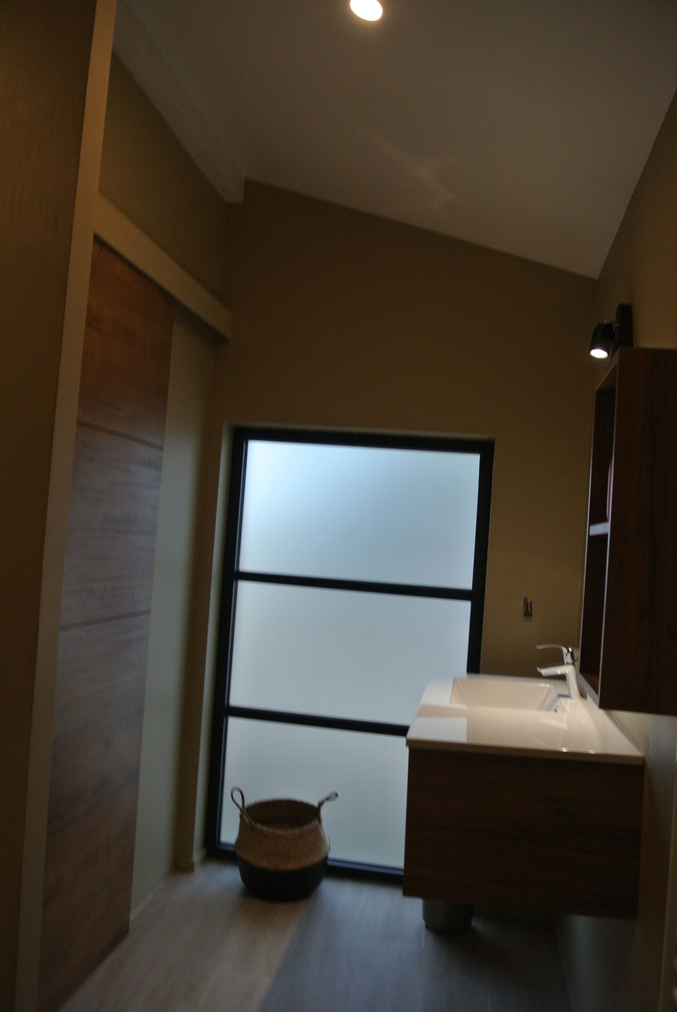 A modern bathroom with a large frosted glass window, a wooden vanity, and a small woven basket on the floor.