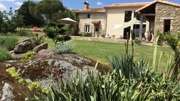 Una casa de piedra con techo de tejas, un jardín con rocas y plantas, y una silla roja bajo una sombrilla.