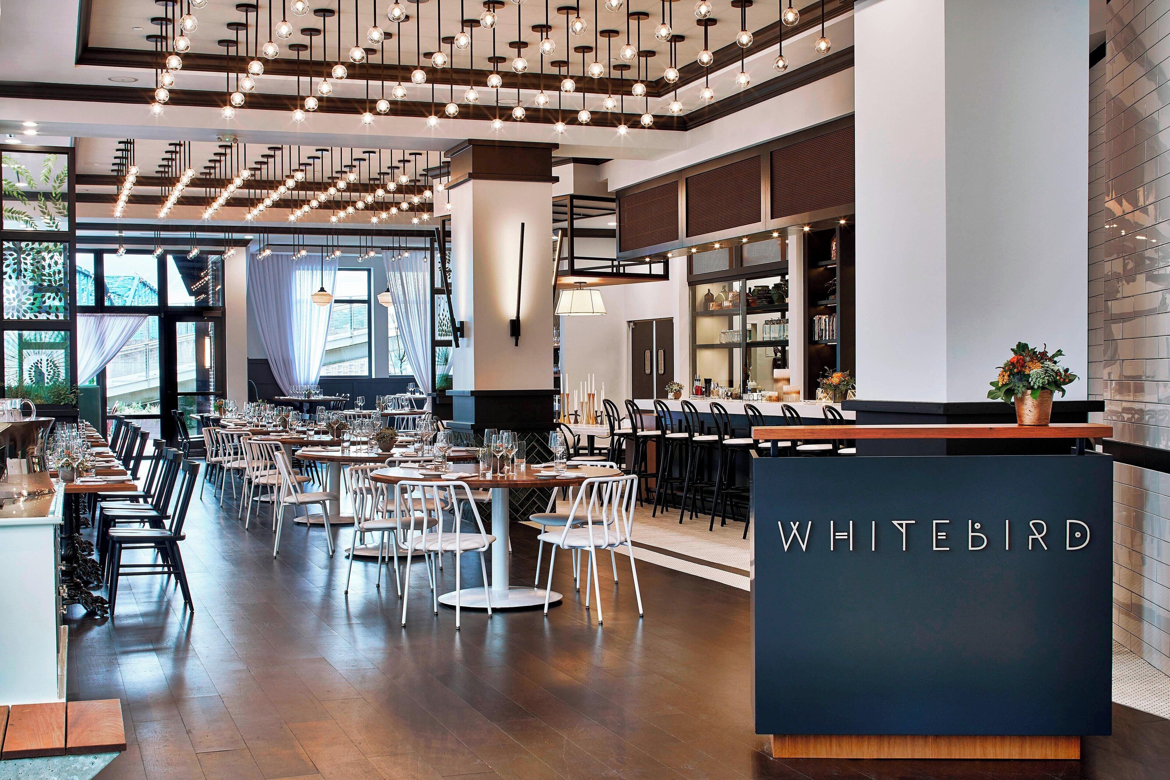 A restaurant interior with a modern design, featuring a long dining area with round tables and chairs, a visible bar area with a counter, and a sign that reads "WHITEBIRD".