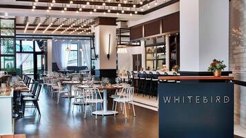 A restaurant interior with a modern design, featuring a long dining area with round tables and chairs, a visible bar area with a counter, and a sign that reads "WHITEBIRD".