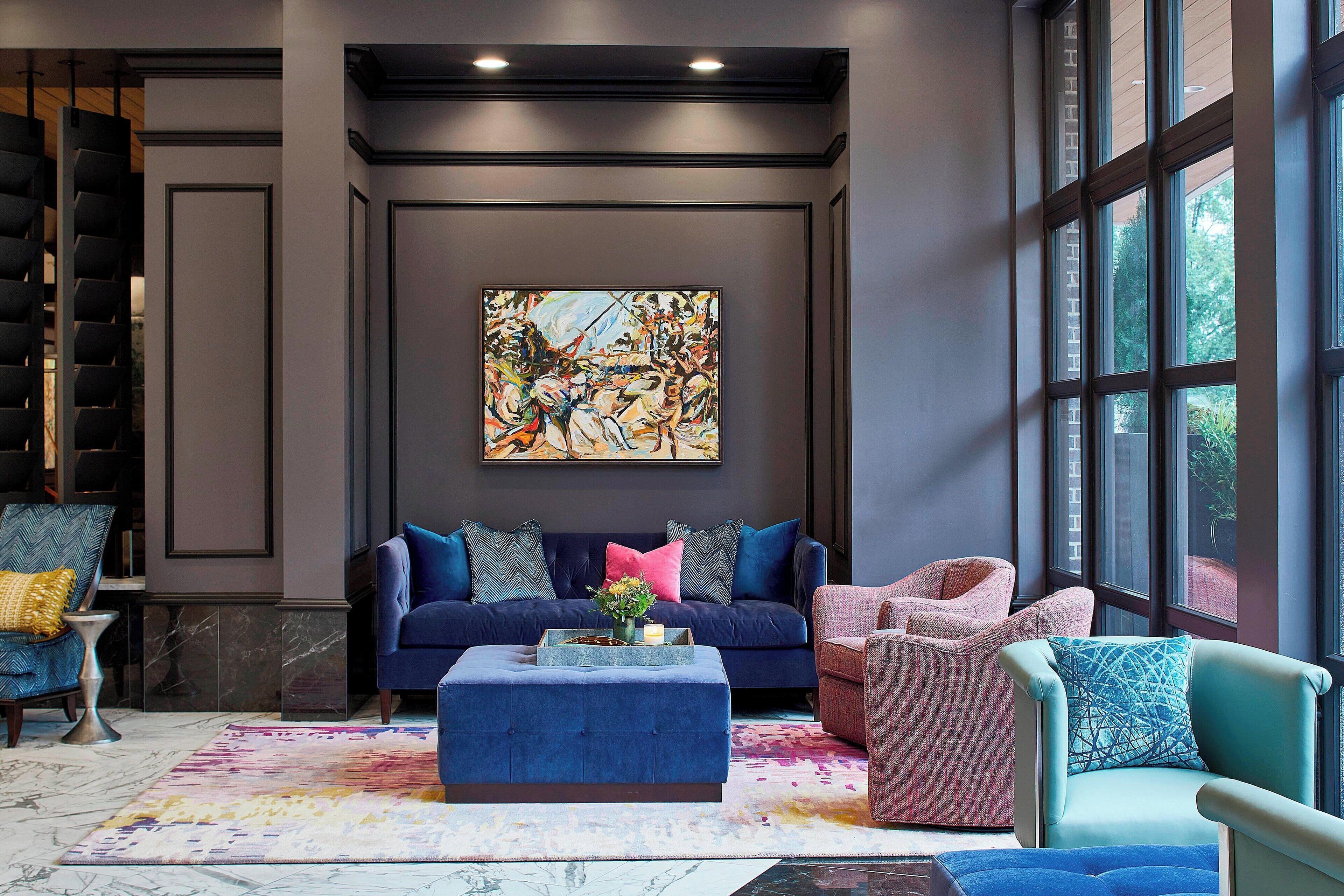 Una sala de estar moderna con un sofá azul, un sillón rosa y un reposapiés azul. Se observa un cuadro abstracto grande en la pared y una ventana que proporciona luz natural.