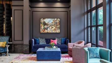 Una sala de estar moderna con un sofá azul, un sillón rosa y un reposapiés azul. Se observa un cuadro abstracto grande en la pared y una ventana que proporciona luz natural.