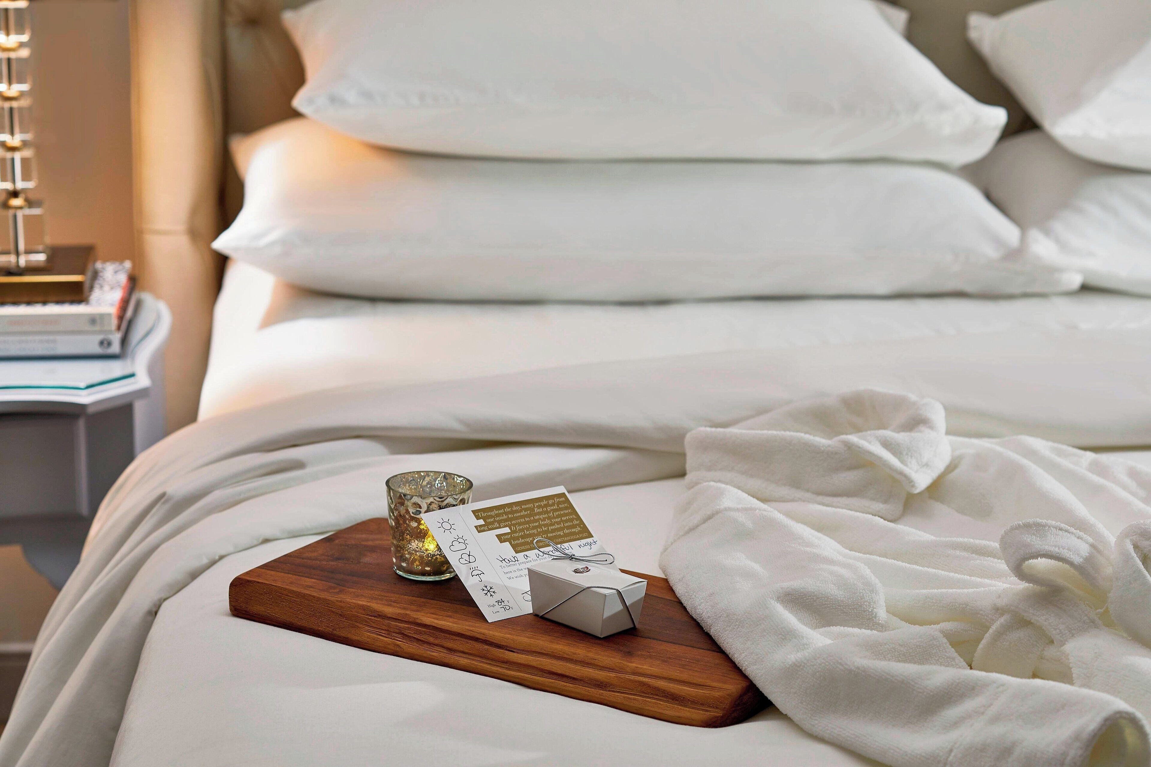 A neatly made bed with white bedding, a wooden tray with a candle, a card, and a small box.