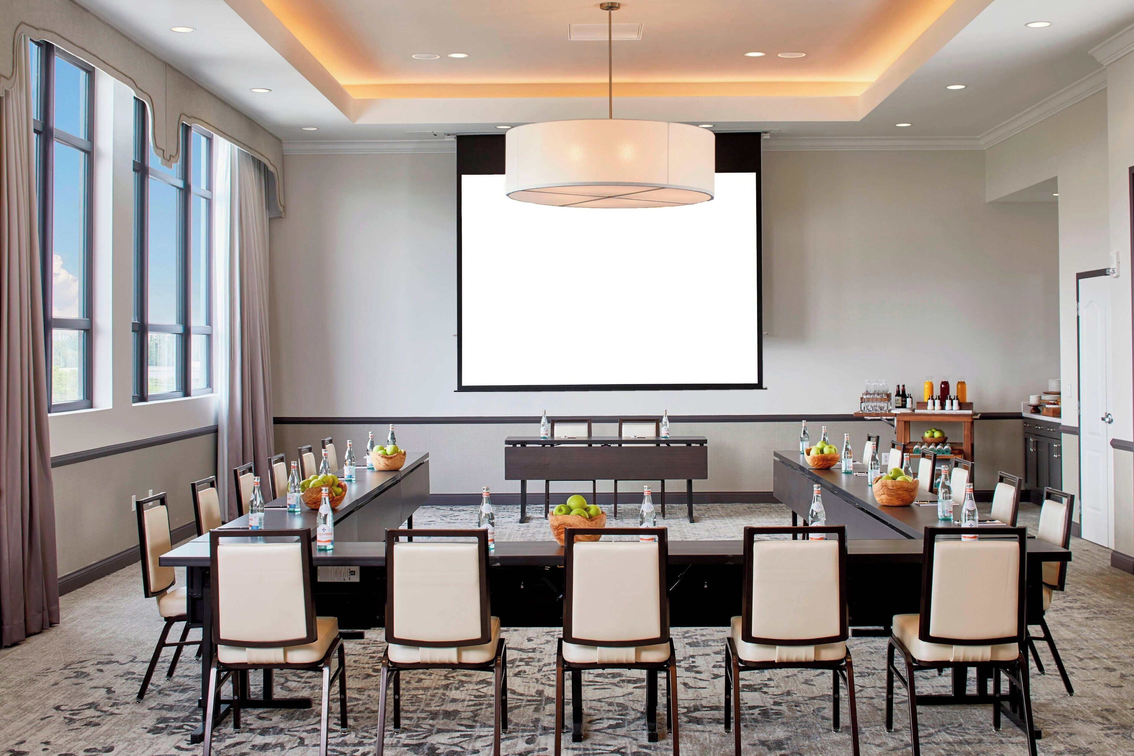 Un salón de conferencias con una larga mesa, sillas, una pantalla de proyector y una zona de buffet con comida y bebidas.